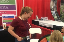 [ai] A young woman wearing a red shirt interacts with children at a booth in an event. The booth has a backdrop featuring posters about equality, justice, and peace.